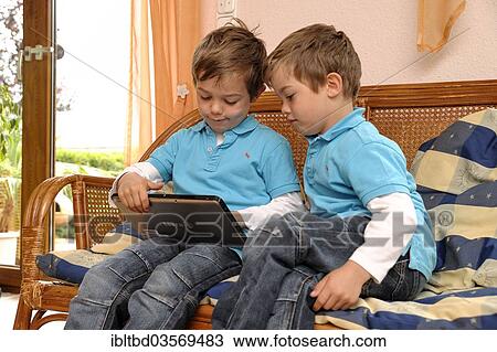 "Two boys, twins, sitting side by side on a bench in a living room playing on a tablet computer, Germany, Europe" View Large Photo Image Stock Image - "Two boys, twins, sitting side by side on a bench in a living room playing on a tablet computer, Germany, Europe". Fotosearch