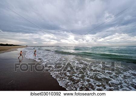 Stock Image - "Two children running into the ocean with clouds providing a threatening mood, Cavallino, Jesolo, Venedig, Italy, Europe". Fotosearch