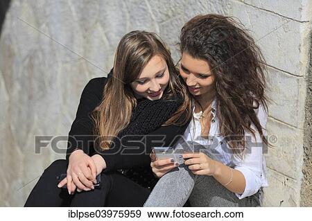 Stock Photo - "Two girlfriends, teenagers, leaning against a wall and reading a text message on the mobile phone, Menton, Provence-Alpes-Cote d'Azur, France, Europe". Fotosearch