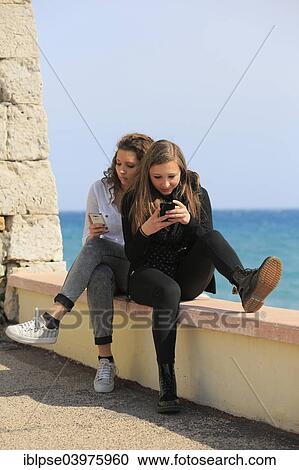 "Two girlfriends, teenagers, sitting on a wall by the sea and writing text messages on their mobile phones, Menton, Provence-Alpes-Cote d'Azur, France, Europe" View Large Photo Image Stock Image - "Two girlfriends, teenagers, sitting on a wall by the sea and writing text messages on their mobile phones, Menton, Provence-Alpes-Cote d'Azur, France, Europe". Fotosearch
