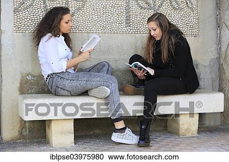 Stock Image - "Two girlfriends, teenagers, sitting on a stone bench, reading books, Menton, Alpes-Maritimes, Provence Alpes, France, Europe". Fotosearch