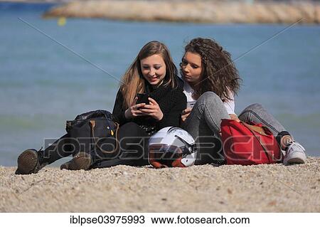 Stock Image - "Two girlfriends, teenagers, sitting on the beach and reading text messages on their mobile phones, Menton, Provence-Alpes-Cote d'Azur, France, Europe". Fotosearch