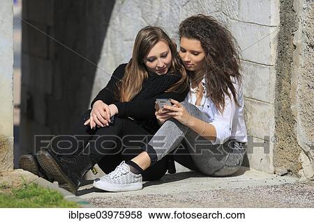 "Two girlfriends, teenagers, sitting on the floor, leaning against a wall and reading a text message on the mobile phone, Menton, Provence-Alpes-Cote d'Azur, France, Europe" View Large Photo Image Stock Photo - "Two girlfriends, teenagers, sitting on the floor, leaning against a wall and reading a text message on the mobile phone, Menton, Provence-Alpes-Cote d'Azur, France, Europe". Fotosearch