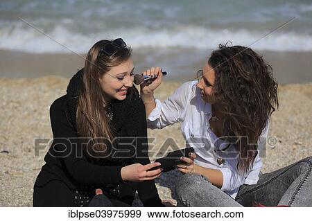Stock Photo - "Two girlfriends, teenagers, sitting on the beach, applying makeup on each other, Menton, Alpes-Maritimes, Provence Alpes, France, Europe". Fotosearch