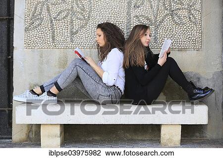 Stock Image - "Two girlfriends, teenagers, sitting back to back on a stone bench, reading books, Menton, Alpes-Maritimes, Provence Alpes, France, Europe". Fotosearch