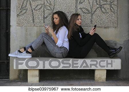 "Two girlfriends, teenagers, sitting back to back on a stone bench and writing text messages on their mobile phones, Menton, Provence-Alpes-Cote d'Azur, France, Europe" View Large Photo Image Stock Photo - "Two girlfriends, teenagers, sitting back to back on a stone bench and writing text messages on their mobile phones, Menton, Provence-Alpes-Cote d'Azur, France, Europe". Fotosearch