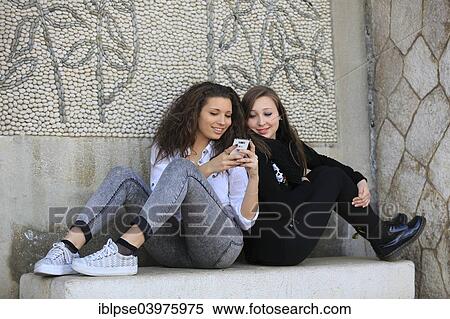 "Two girlfriends, teenagers, sitting back to back on a stone bench and reading a text message on a mobile phone, Menton, Provence-Alpes-Cote d'Azur, France, Europe" View Large Photo Image Stock Photography - "Two girlfriends, teenagers, sitting back to back on a stone bench and reading a text message on a mobile phone, Menton, Provence-Alpes-Cote d'Azur, France, Europe". Fotosearch
