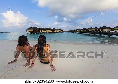 Stock Image - "Two girls, about 15 and 19 years old sitting on the beach, lagoon, Paradise Island at the back, Lankanfinolhu, North Male Atoll, Republic of Maldives, Indian Ocean, Asia". Fotosearch