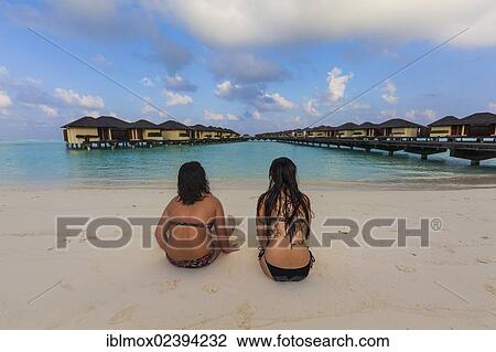 Stock Image - "Two girls, about 15 and 19 years old sitting on the beach, lagoon, Paradise Island at the back, Lankanfinolhu, North Male Atoll, Republic of Maldives, Indian Ocean, Asia". Fotosearch