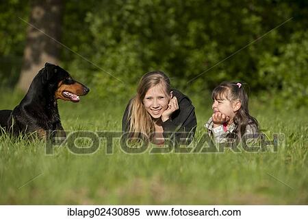 Stock Photography - Two girls lying with a Doberman on a meadow. Fotosearch