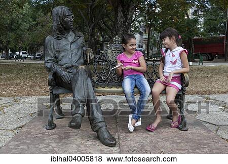 Stock Photo - "Two girls sitting on a bench next to a bronze sculpture of John Lennon, Havana, Cuba, North America". Fotosearch