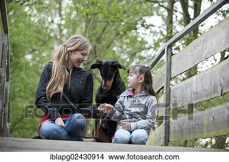 Two girls sitting with a Doberman on a bridge View Large Photo Image Picture - Two girls sitting with a Doberman on a bridge. Fotosearch