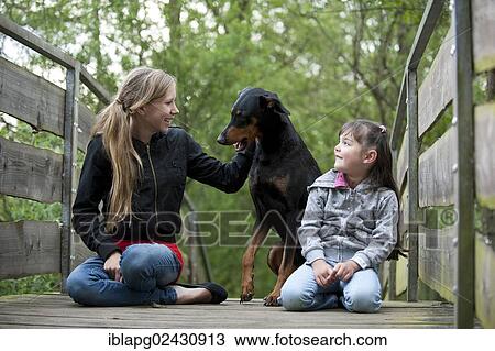 Two girls sitting with a Doberman on a bridge View Large Photo Image Stock Image - Two girls sitting with a Doberman on a bridge. Fotosearch