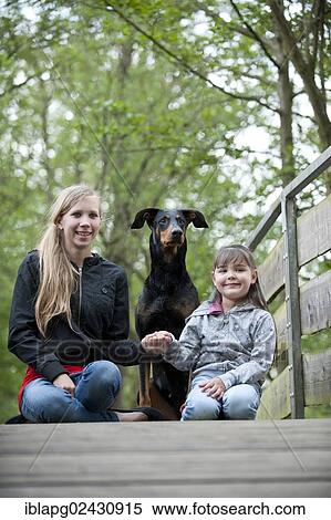 Two girls sitting with a Doberman on a bridge View Large Photo Image Stock Photography - Two girls sitting with a Doberman on a bridge. Fotosearch