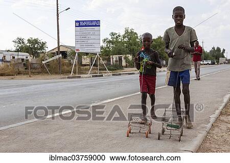 Stock Image - "Two Himba boys playing with their self-made toy cars by the roadside, Opuwo, Kaokoland, Kunene, Namibia, Africa". Fotosearch