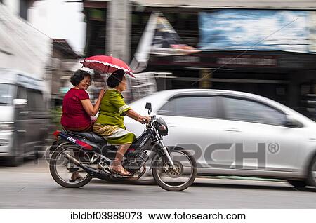 Stock Image - "Two women on a motorcycle, with red parasol, Chiang Mai, Northern Thailand, Thailand, Asia". Fotosearch