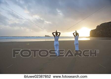 Stock Image - Two women relaxing by doing yoga on the beach at sunset. Fotosearch