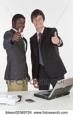 Picture - Two young businessmen of different races with a laptop and making a thumbs-up gesture. Fotosearch