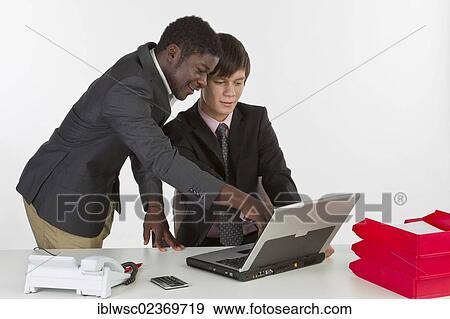 Stock Photo - Two young businessmen of different races working on a laptop and discussing with each other. Fotosearch