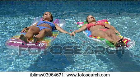 Stock Image - "Two young women, friends, holding hands, relaxing on air mattress in pool, Puerto del Carmen, Lanzarote, Canary Islands, Spain, Europe". Fotosearch