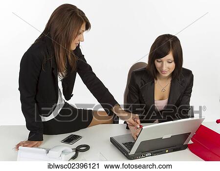 "Two young women sitting at a desk with a laptop, discussing" View Large Photo Image Stock Image - "Two young women sitting at a desk with a laptop, discussing". Fotosearch