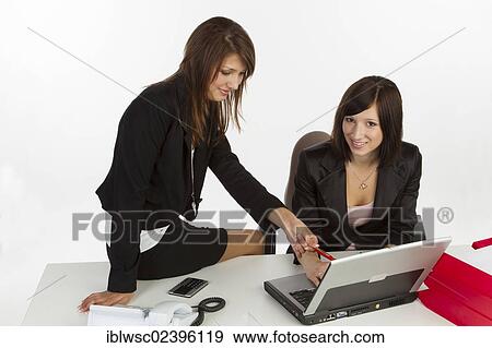 "Two young women sitting at a desk with a laptop, discussing" View Large Photo Image Stock Photo - "Two young women sitting at a desk with a laptop, discussing". Fotosearch