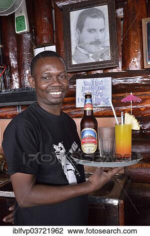 Stock Image - "Waiter with Kilimanjaro beer and a cocktail in front of a picture of Freddy Mercury, the late singer of the rock band Queen who was born in Zanzibar, Mercury's Restaurant, Stone Town, Zanzibar City, Zanzibar, Tanzania, Africa". Fotosearch