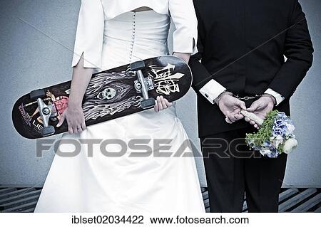 Stock Image - "Wedding couple seen from behind, the bride is holding a skateboard, the groom is wearing handcuffs and holding a bridal bouquet". Fotosearch