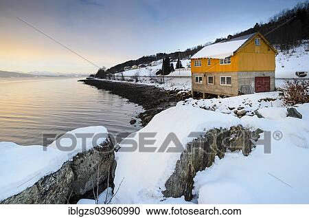 Stock Image - "Winter atmosphere at sunrise on the fjord, snow-covered rocks at the front, typical Norwegian wooden house and coastal road, near Alesund, Norway, Europe". Fotosearch