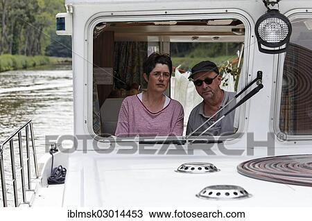 Stock Image - "Woman and man at the wheel of a Penichette, a houseboat on the Canal des Vosges, formerly Canal de l'Est, Fontenoy-le-Chateau, Epinal, Lorraine, Departement Vosges, France, Europe". Fotosearch