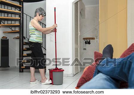 Stock Image - "Woman cleaning while a man is lying on the couch, Germany, Europe". Fotosearch
