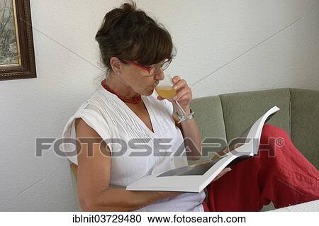 Stock Image - "Woman, mid 60s, drinking a glass of wine while reading". Fotosearch