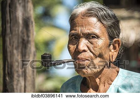 Stock Image - "Woman of the Chin people, ethnic minority, with a traditional facial tattoo smoking a pipe, the last of their kind, portrait, Rakhine State, Myanmar, Asia". Fotosearch
