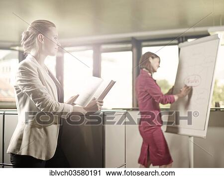 Stock Image - "Woman reading a book and looking towards her colleague who is writing on a flip chart, Innsbruck, Tyrol, Austria, Europe". Fotosearch