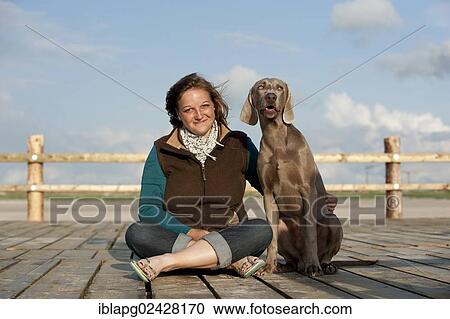 Stock Image - Woman sitting with a Weimaraner on a jetty. Fotosearch