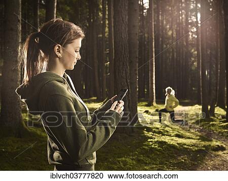 "Woman standing in the forest checking her smartphone, another woman doing stretching exercises at back, Tyrol, Austria, Europe" View Large Photo Image Stock Image - "Woman standing in the forest checking her smartphone, another woman doing stretching exercises at back, Tyrol, Austria, Europe". Fotosearch