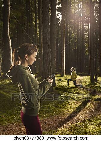"Woman standing in the forest checking her smartphone, another woman doing stretching exercises at back, Tyrol, Austria, Europe" View Large Photo Image Stock Photo - "Woman standing in the forest checking her smartphone, another woman doing stretching exercises at back, Tyrol, Austria, Europe". Fotosearch