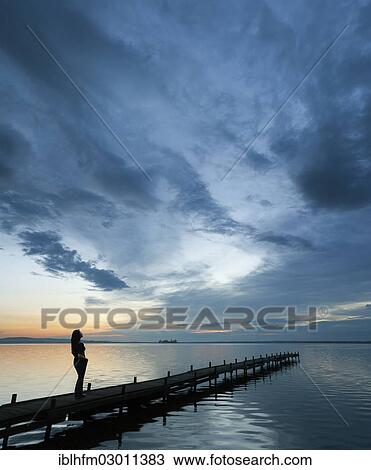 Stock Image - "Woman standing on a wooden pier on the lake admiring the beautiful night sky, Steinhuder Meer, Steinhude, Lower Saxony, Germany, Europe". Fotosearch