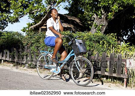 Stock Image - "Woman talking on a mobile phone while riding a bicycle, La Digue Island, Seychelles, Africa, Indian Ocean, Africa". Fotosearch
