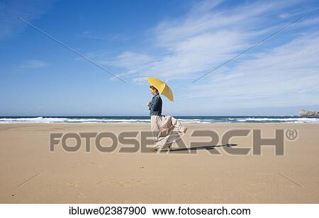 Stock Image - Woman walking with an umbrella on a beach. Fotosearch