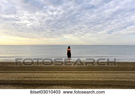 Stock Image - "Woman wearing a black dress standing by the sea, moody clouds, Cavallino, Jesolo, Venedig, Italy, Europe". Fotosearch