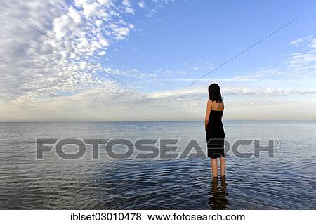Stock Photo - "Woman wearing a black dress standing in the water of the sea, moody clouds, Cavallino, Jesolo, Venedig, Italy, Europe". Fotosearch