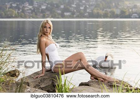 Stock Photograph - "Woman wearing a white swimsuit sitting on a rock on the lake shore, next to a swan". Fotosearch