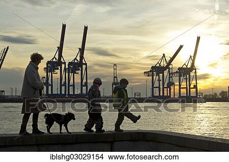 Picture - "Woman with a dog and two children walking along a wall in front of gantry cranes, Container Terminal Burchardkai, Hamburg-Port, Hamburg, Hamburg, Germany, Europe". Fotosearch