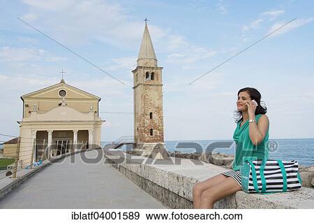 Stock Photo - "Woman with cell phone sitting on the wall in front of the church Madonna dell 'Angelo, Adriatic, Caorle, Veneto Region, Italy, Europe". Fotosearch