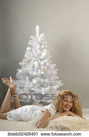 Stock Image - "Women, 25-35 years old, in a white dress, is lying on a bearskin rug in front of a white Christmas tree". Fotosearch