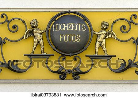 Stock Image - "Wrought iron sign for """"Hochzeitsfotos"""" or wedding photos with cherubs on a photo shop, Murnau, Upper Bavaria, Bavaria, Germany, Europe". Fotosearch