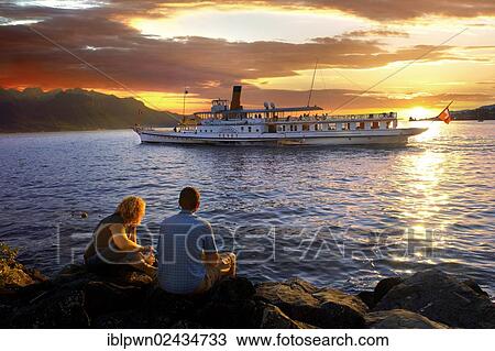 Stock Image - "Youg couple watching a ferry at sunset on Lac Leman, Lake Geneva, Montreux, Switzerland, Europe". Fotosearch