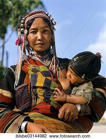 Stock Image - "Young Akha woman breastfeeding baby, with traditional clothing and headgear, Chiang Rai Province, Northern Thailand, Thailand, Asia". Fotosearch