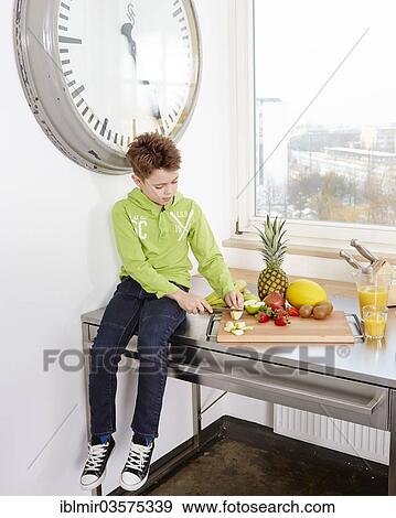 Stock Photo - "Young boy sitting on a kitchen worktop, cutting various kinds of fruit on a cutting board, Germany, Europe". Fotosearch